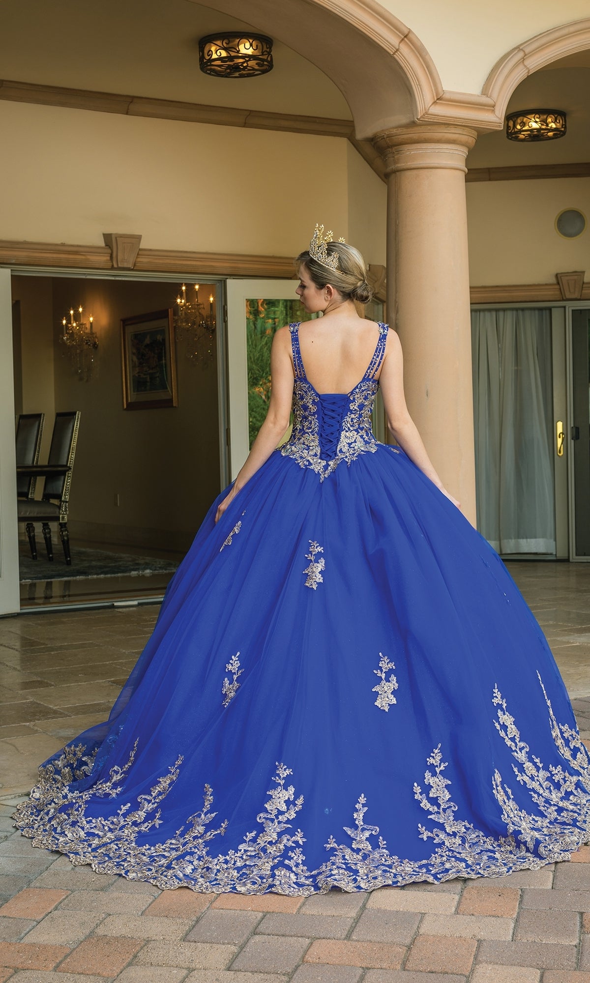 A woman wearing the Royal Blue Quinceanera Dress 1593, featuring gold embroidery and a crown, stands with her back to the camera before a columned building.